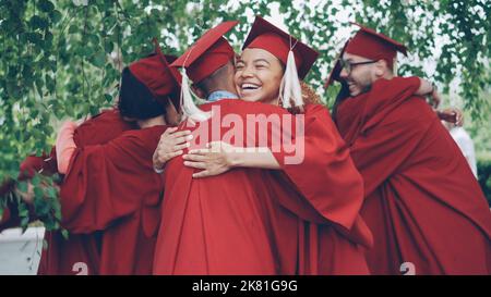 Fröhliche Freunde, die Studenten in Kleidern und Leichenbrettern graduieren, gratulieren sich nach der Abschlussfeier, umarmen und lachen. Menschen- und Bildungskonzept. Stockfoto