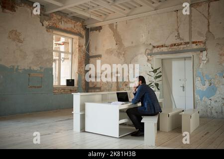 Porträt eines jungen Geschäftsmannes, der in einem leeren Raum mit unvollendeter Restaurierung und Denkweise sitzt. Ausfall des Geschäftsstarts Stockfoto