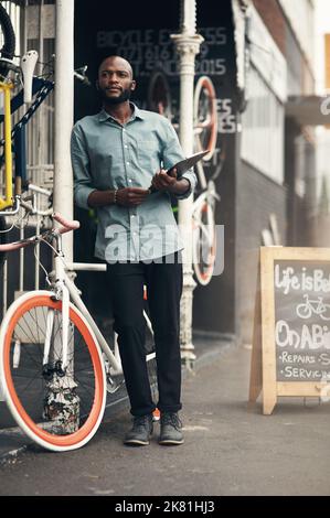 Wo ist diese Lieferung? Die ganze Aufnahme eines hübschen jungen Mannes, der vor seinem Fahrradladen steht und besinnlich aussieht, während er ein Klemmbrett hält Stockfoto