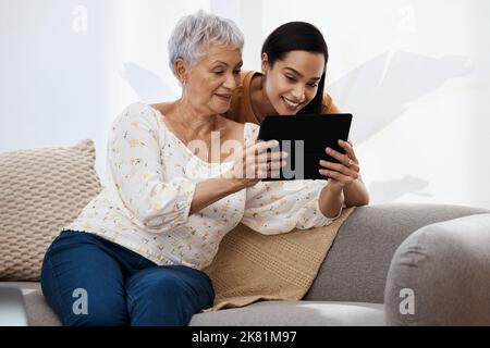 Eine ältere Frau, die mit ihrer Tochter zu Hause auf dem Sofa ein digitales Tablet benutzt, muss es der Technologie übergeben, um uns zusammenzubringen. Stockfoto