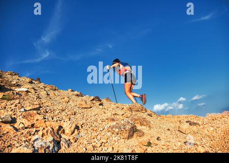 Junge Frau bergauf auf auf felsigen Hang in Berg schiebt mit Stöcken Stockfoto