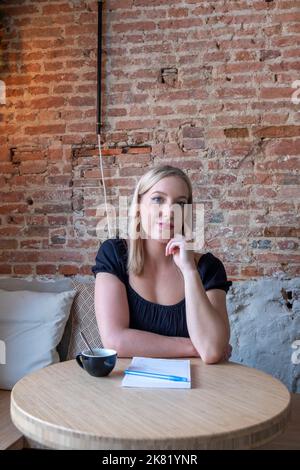 Verträumte junge Frau, die Zukunftspläne und Ziele in einem eigenen Tagebuch niederschreibt, die sich in einer gemütlichen Cafeteria mit einer Tasse leckeren Cappuccino ausruht.lächelndes Hipster-Mädchen, das beim Schreiben auf einem Notizblock eine gute Idee macht. Hochwertige Fotos Stockfoto