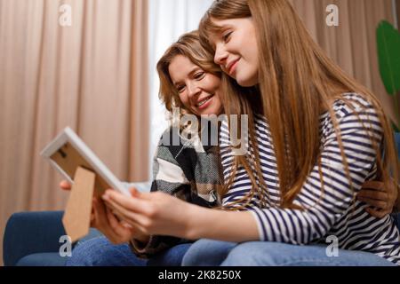 Die junge Frau, die ihre Mutter besucht, sitzt auf dem Sofa und schaut auf ein Bild in einem Rahmen. Mutter und Tochter beim Betrachen alter Fotos Stockfoto
