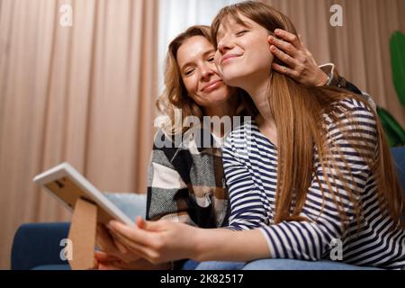 Glückliche Mutter und Tochter umarmen sich, während sie auf dem Sofa sitzen und auf ein gerahmtes Foto schauen Stockfoto