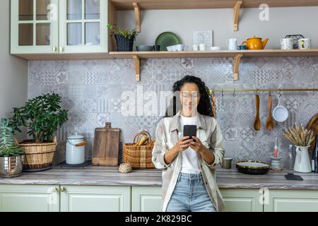 Hispanische Frau benutzt zu Hause in der Küche ein Mobiltelefon, lächelt und freut sich, liest Nachrichten, durchstösst Webseiten, kommuniziert online mit Freunden und Dating. Stockfoto