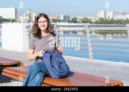 Ein lächelndes Studentenmädchen mit Brille, öffnete ihren Rucksack, suchte nach etwas in ihrer Tasche, saß draußen auf einer Bank auf einer Stadtstraße. Ein Teenager s Stockfoto