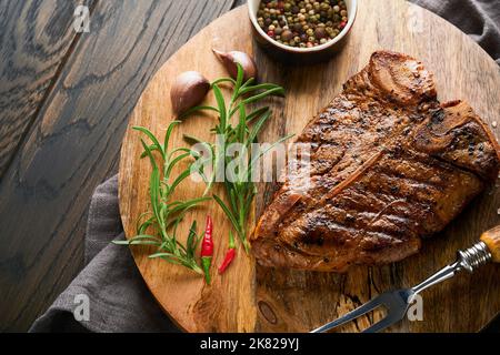 Rohes Steak, T-Bone oder Porterhouse. Steak-T-Bone oder Porterhouse in Sauce mit Gewürzen, Salz und Rosmarin auf einem schwarzen Keramikplatte auf Betonboden. Stockfoto