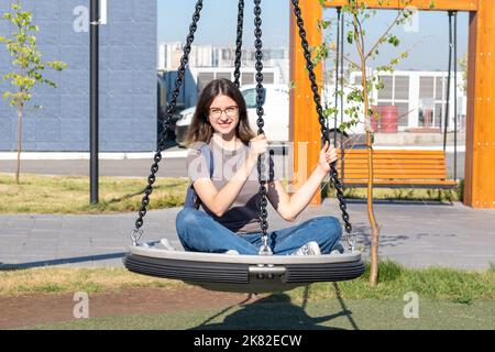 Spanisch lächelnde Mädchen Student in Brille Jeans schwingen auf einer Schaukel im Park. Das Mädchen ruht sich nach dem Studium aus. Sommer-Unterhaltung in der Stadt pa Stockfoto