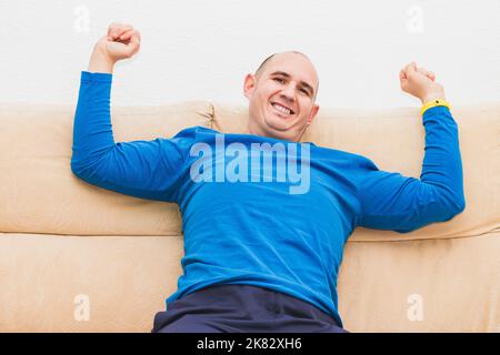 Ein kahlköpfiger Kaukasusmann, der in einem blauen Langarm-T-Shirt gekleidet ist, streckt sich auf einem Sofa aus. Stockfoto