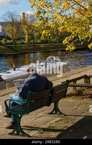 Ein erwachsener Mann, der sich beim Lesen einer Zeitung am Fluss Ouse am Dame Judi Dench Walk, York, North Yorkshire, Großbritannien, entspannt. Stockfoto