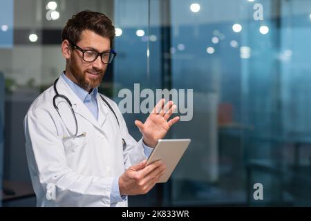 Fröhlicher Oberarzt im medizinischen Mantel mit Tablet-Computer für Videoanruf und Online-Beratung mit Patient, Mann mit Bart und Brille in medizinischen Mantel arbeiten innerhalb der Klinik. Stockfoto