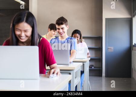 Große Gruppe von multiethnischen lächelnden Studenten, die an Laptops arbeiten, während sie einer Vorlesung im Klassenzimmer zuhören. Junge fröhliche Menschen lernen bei Stockfoto