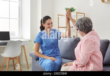 Fröhliche, positive Krankenschwester, die ihrer älteren Patientin während des Besuchs hohe fünf Punkte gibt. Stockfoto