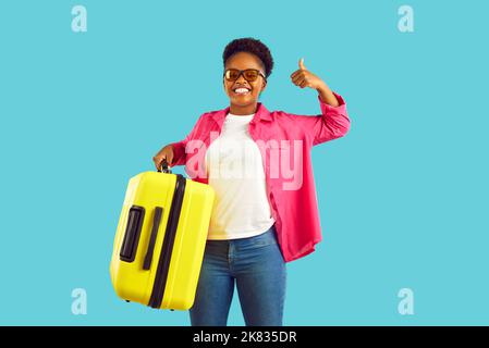 Eine afroamerikanische Frau in einer Sonnenbrille zeigt den Daumen mit einem gelben Koffer auf türkisfarbenem Hintergrund. Stockfoto