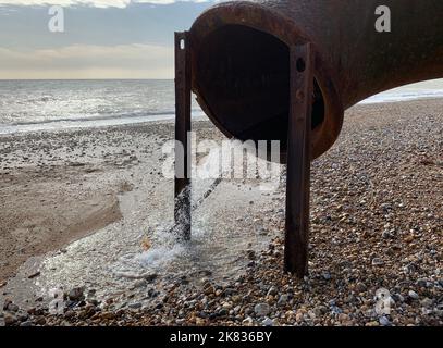 Nahaufnahme eines großen rostigen Eisenabflussrohres mit Wasser, das am Strand der Südküste Großbritanniens herausströmt. Stockfoto