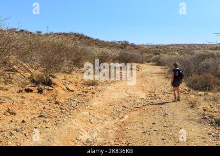 Oanob, Namibia - 28. September 2018: Frau auf einem Pfad im Oanob Resort. Namibische Landschaft entlang des Schotterweges. Roter Boden und afrikanische Vegetation aroun Stockfoto