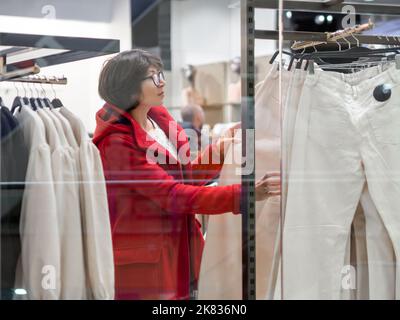 Die Frau wählt im Bekleidungsgeschäft Hosen aus. Legere Hose an Kleiderbügeln. Blick durch transparentes Schaufenster. Einkaufen im Einkaufszentrum. Einfache Kleidung. Stockfoto