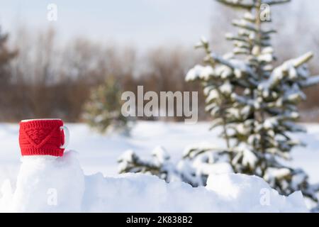 Ein roter Becher mit einem heißen Getränk bei einer Schneeverwehung an einem frostigen sonnigen Wintertag im Wald vor dem Hintergrund schneebedeckter Tannenbäume. Landschaft. Wählen Sie Stockfoto