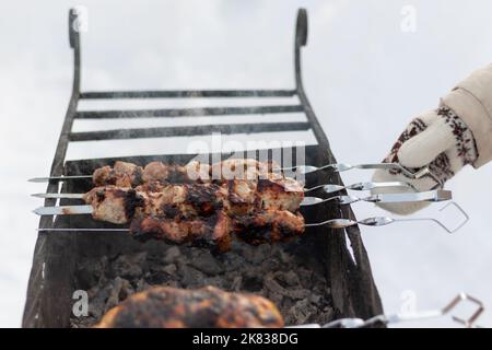 Eine Frauenhand in einem Fäustling fritiert an einem sonnigen Wintertag einen Grill auf dem Grill in der Natur. Selektiver Fokus. Nahaufnahme Stockfoto