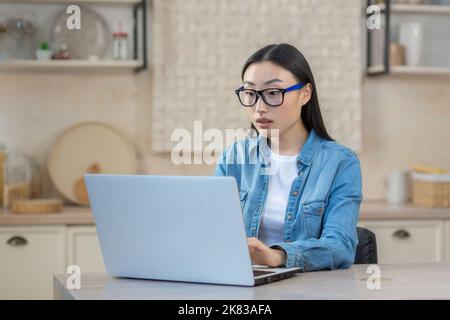 Fokussierte und seriöse junge asiatische Frau freiberufliche Arbeit zu Hause auf Laptop. Am Tisch sitzen, online arbeiten. Stockfoto