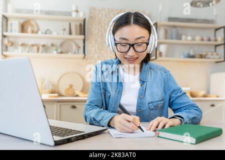 Online-Schulungen in Quarantäne. Junge schöne asiatische Student Mädchen sitzen am Tisch zu Hause tragen weiße Kopfhörer und mit einem Laptop. Er studiert aus der Ferne. Schreibt in ein Notizbuch. Stockfoto