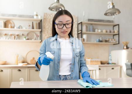 Zimmerreinigung. Junge schöne asiatische Frau arbeitet als Reinigungskraft. Reinigt das Haus. Wischt den Tisch mit einem Tuch in Gummihandschuhen, sprüht die Oberfläche mit einem Spray. Stockfoto