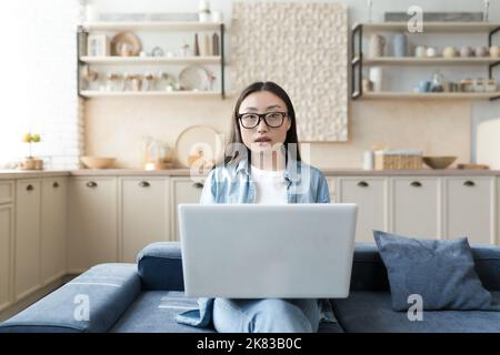 Porträt einer jungen schönen asiatischen Bloggerin. Zu Hause auf der Couch sitzend, einen weißen Laptop auf dem Schoß haltend, aufmerksam auf die Kamera schauend. Stockfoto