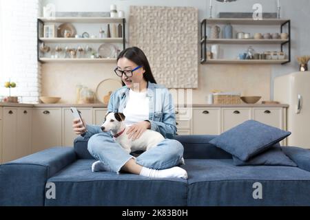 Eine glückliche junge, schöne asiatische Frau in Brille spricht bei einem Videoanruf, hält ein Telefon und ihren Hund in den Händen und lächelt die Kamera an. Sitzen zu Hause auf dem Sofa in der Lotusposition. Stockfoto
