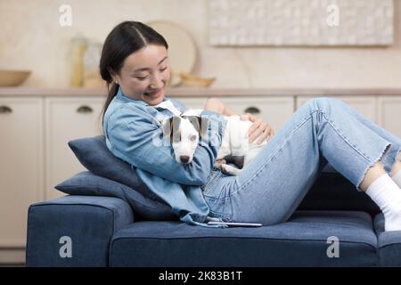 Eine lächelnde junge, schöne asiatische Frau sitzt zu Hause auf dem Sofa und hält einen kleinen Hund in den Händen. Seine Freizeit verbringt er mit seinem Haustier, spielt, ruht sich aus Stockfoto