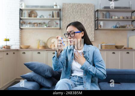 Allergischer Angriff. Asthma. Junge asiatische Frau, die zu Hause auf dem Sofa sitzt und Inhalator verwendet. Hat einen Asthmaanfall, behandelt Allergien, hat Atembeschwerden. Stockfoto