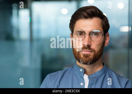 Nahaufnahme eines ernsthaften und nachdenklichen, reifen Geschäftsmannes mit Bart, Mann im Hemd und Brille, der die Kamera anschaut und in einem modernen Bürogebäude starrend arbeitet. Stockfoto