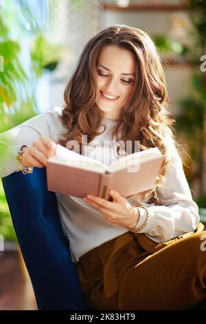 Green Home. Lächelnd stilvolle Hausfrau mittleren Alters mit langen welligen Haaren mit Buch in grünen Hosen und graue Bluse im modernen Wohnzimmer an sonnigen Tagen. Stockfoto