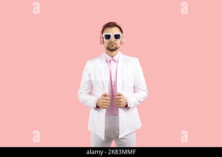 Modischer selbstbewusster Mann in einem weißen klassischen Anzug und einem rosa Hemd in Sonnenbrille mit Kopfhörern auf einem rosa Hintergrund. Stockfoto