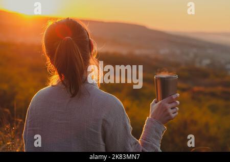 Rückansicht einer Frau, die bei Sonnenuntergang eine heiße Tasse Tee auf dem Hügel genießt Stockfoto