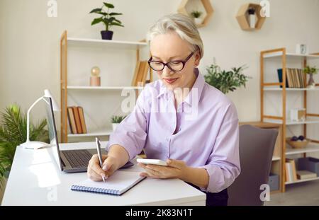 Reife Geschäftsfrau, die am Schreibtisch sitzt, Mobiltelefon hält und im Notebook schreibt Stockfoto