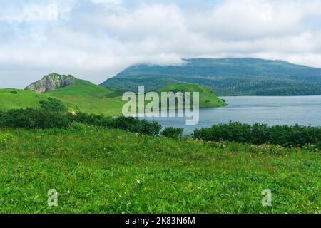 Natürliche Landschaft der Insel Kunashir mit grasbewachsenen Hügeln, vulkanischen Felsen, Vulkan in den Wolken und einem Tal mit einer Lagune Stockfoto