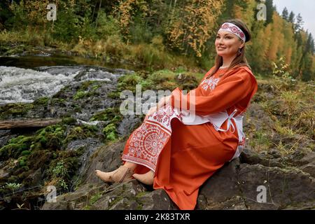 Eine Frau in Nationaltracht am Kivach Wasserfall in Karelien im Herbst. Die Republik Karelien auf der Kola-Halbinsel ist eine der nördlichen Regionen Russlands, die an Finnland grenzt. Diese dünn besiedelten Orte haben eine reiche Geschichte und haben viele natürliche Ecken in ihrer ursprünglichen Form erhalten. Stockfoto