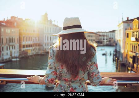 Von hinten gesehen eine moderne Reisende Frau in Blumenkleid mit Hut bei der Besichtigung der Accademia-Brücke in Venedig, Italien. Stockfoto