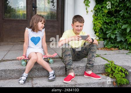 Kinder im Freien sitzen auf der Treppe und sehen sich Videos am Telefon an. Soziales Netzwerk Stockfoto