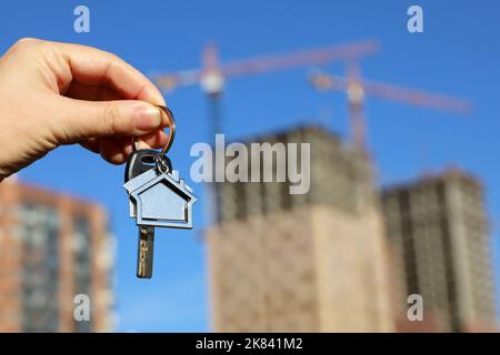 Immobilienmakler, Schlüsselanhänger in Form eines Hauses und Schlüssel in weiblicher Hand auf dem Hintergrund von Baukräne und Neubauten. Byung Wohnung oder Miete Stockfoto