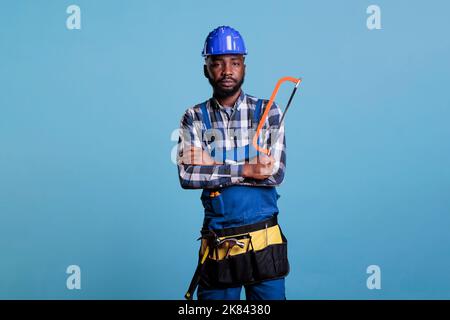 Seriöse Bauarbeiter halten Säge Blick auf Kamera isoliert auf blauem Hintergrund. Zimmermann hält seine Arbeitswerkzeuge tragen Uniform und Schutzhelm, Studio Schuss. Stockfoto