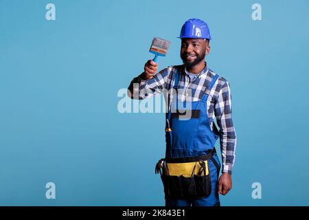 Professioneller Hausmaler malt neues Haus hält Pinsel trägt Schutzhelm und Overalls. Reparaturman glücklich macht Innenrenovierung isoliert auf blauem Hintergrund. Stockfoto