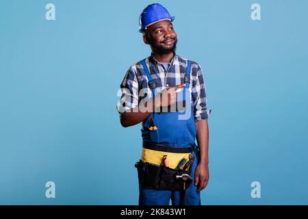 Porträt eines glücklichen Bauarbeiters bei der Arbeit, der einen harten Hut trägt und seinen Finger zur Seite zeigt. Elektriker trägt Gürtel mit Werkzeugen im Studio vor blauem Hintergrund aufgenommen. Stockfoto