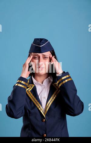 Erschöpfte Stewardess mit Migränesymptomen, Schläfen mit Fingern berühren, mit Grimasse auf die Kamera schauen. Kranker Flugbegleiter in Fluguniform, der unter Kopfschmerzen leidet Stockfoto