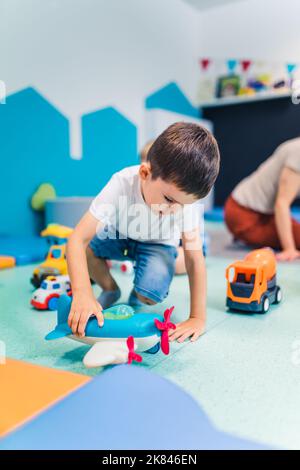 Kaukasischer kleiner Junge, der im Kindergarten mit einem Plastikflugzeug spielt, vertikale Aufnahme. Hochwertige Fotos Stockfoto