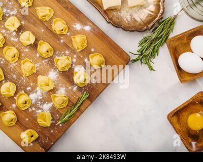 Auf einem bemehlten Holzschneidebrett, rohe hausgemachte Knödel mit Fleischfüllung, Küchenutensilien und Zutaten auf weißem Hintergrund. Low-Angle-Ansicht. R Stockfoto