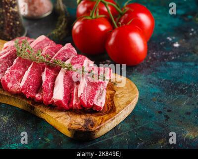 Appetitliche rohe Steaks aus Bio-Fleisch vom Bauernhof auf einem Holzschneidebrett, reife frische Tomaten und Gewürze auf dunklem Marmorboden. Wunderschön Stockfoto