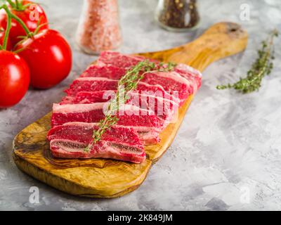 Nahaufnahme. Rohe Fleischsteaks mit einem Rosmarinzweig auf einem Holzschneidebrett, Tomaten, Gewürzen und Gewürzen auf weißem Hintergrund. Rezepte für Resta Stockfoto