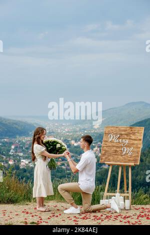 Angebot in den Bergen. Ein Kerl legt einen Ring auf seine Braut, während er auf seinem Knie steht. Glückliches Paar. Heiraten Sie mich. Viel Glück mit dem Vorschlag. Stockfoto