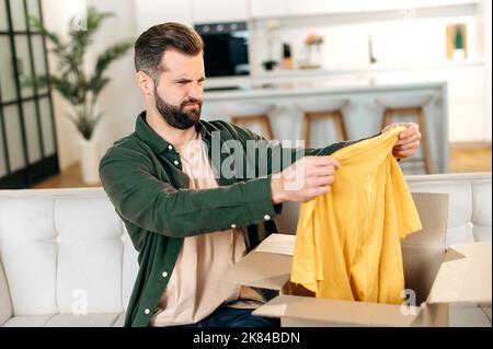 Trauriger kaukasischer Mann in legerer Kleidung, sitzt auf einem Sofa im Wohnzimmer, packt sein Paket aus, nimmt Kleidung aus dem Karton, schaut auf sie enttäuscht, unzufrieden, ungerechtfertigte Erwartungen Stockfoto